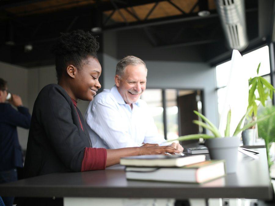 A young woman and an older man smiling as they work together on a computer at a desk in a bright, modern office.
