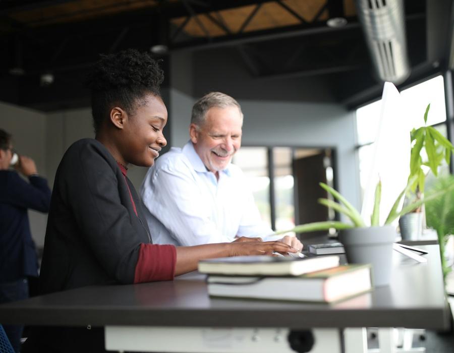 A young woman and an older man smiling as they work together on a computer at a desk in a bright, modern office.