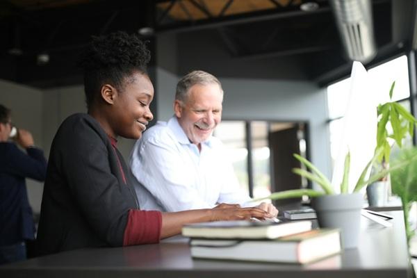 A young woman and an older man smiling as they work together on a computer at a desk in a bright, modern office.