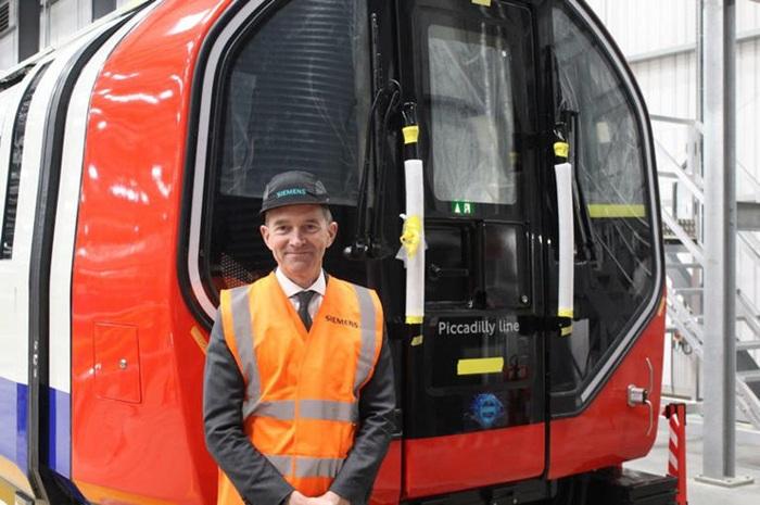 Man in an orange high-vis vest stood in a factory in front of a London Underground train.