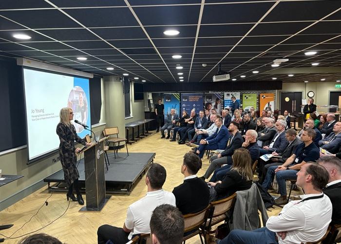 A woman stands at a podium speaking at a business event, while a large audience of mostly seated adults listens attentively in a conference room.