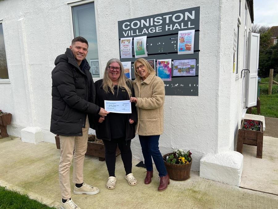 Three people stand outside Coniston Village Hall, smiling while holding a cheque in front of a noticeboard.