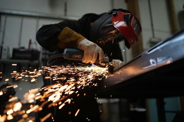 Worker wearing protective gloves and a face shield uses an angle grinder on a metal beam, producing bright sparks in a workshop.