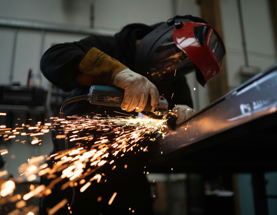 Worker wearing protective gloves and a face shield uses an angle grinder on a metal beam, producing bright sparks in a workshop.