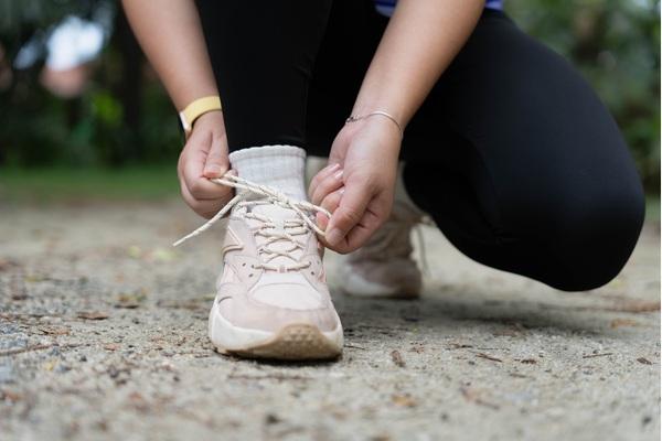 A person outdoors in athletic wear, kneeling on a gravel path to tie the laces of their cream-colored sneaker.