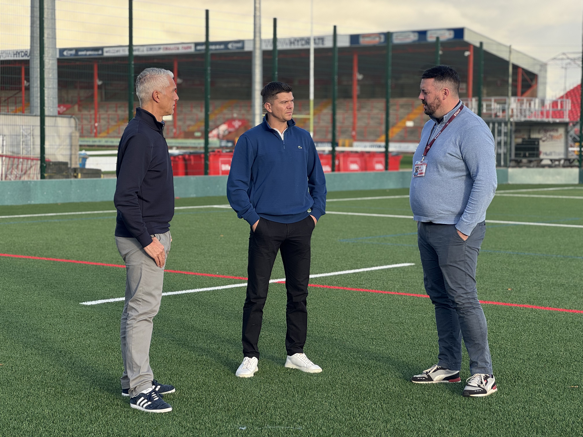 Mayor Luke Campbell and two other men standing and talking on an outdoor sports field with artificial turf.