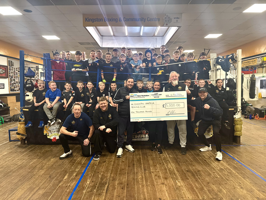 Large group of children and adult coaches posing inside a boxing gym at Kingston Amateur Boxing Club, gathered around a boxing ring while holding a £5,000 donation cheque, smiling and celebrating.