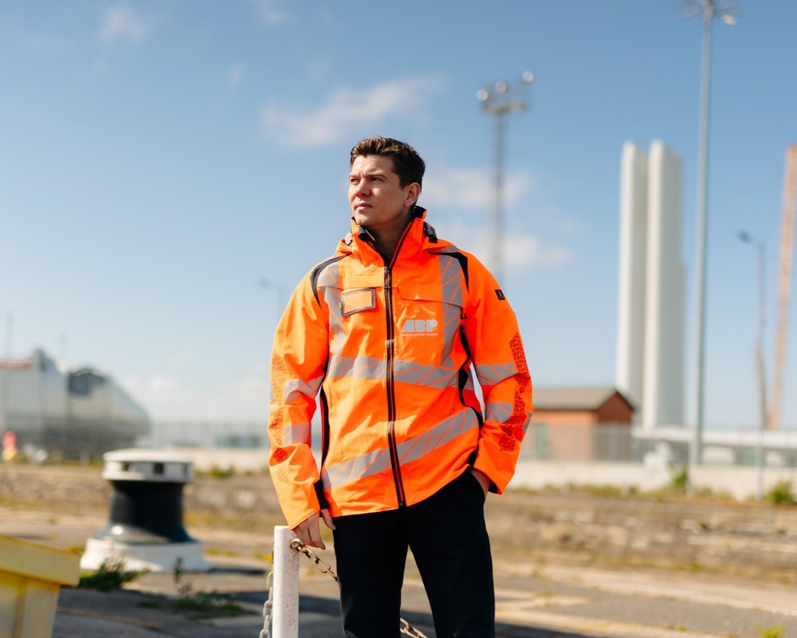 Mayor Luke wearing a bright orange high-visibility safety jacket stands outdoors at an industrial site, looking into the distance under a clear blue sky.