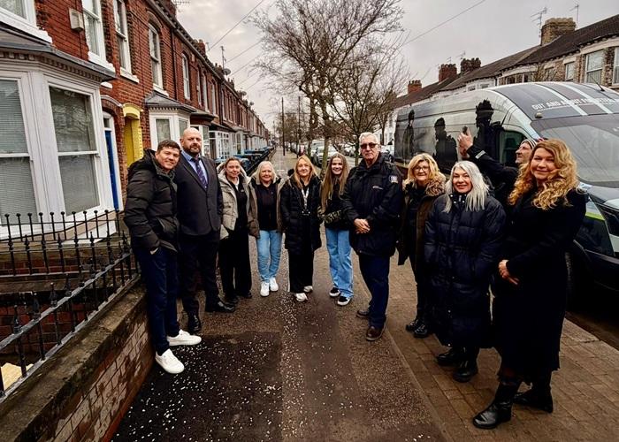 A group of people stand together, talking and smiling, on a residential street lined with terraced houses.
