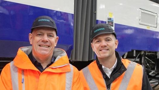 Two men wearing orange high-visibility jackets and Siemens-branded caps stand side by side in front of a blue-and-white train, smiling at the camera.