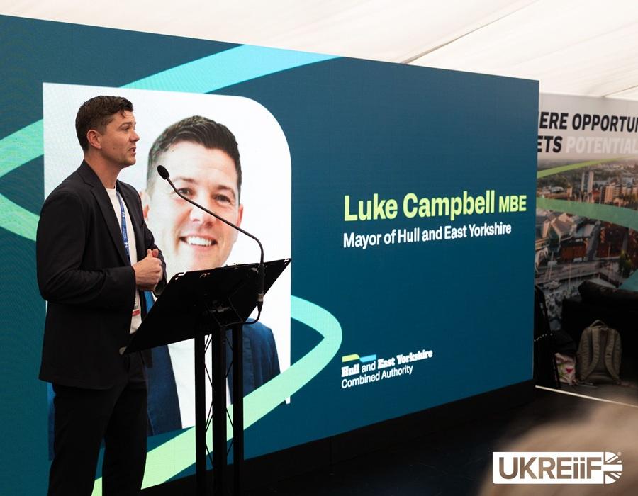 Mayor Luke Campbell speaking at a podium in front of a large presentation screen at a business event.