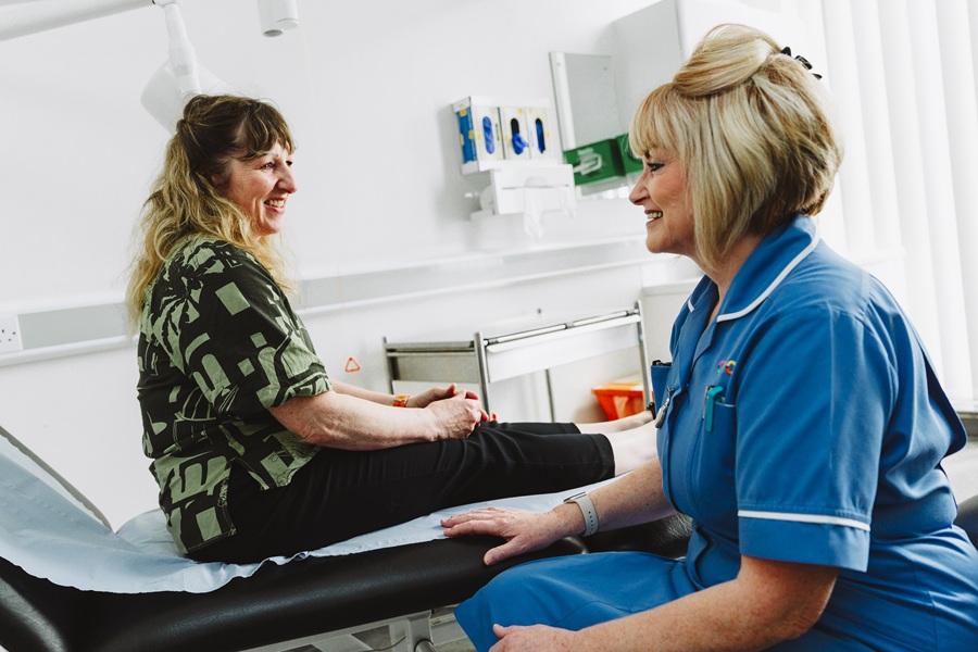A nurse speaks with a smiling patient seated on an examination bed during a healthcare appointment.