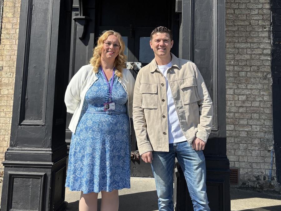 Two people stand side by side outside a dark-painted brick building on a sunny day.