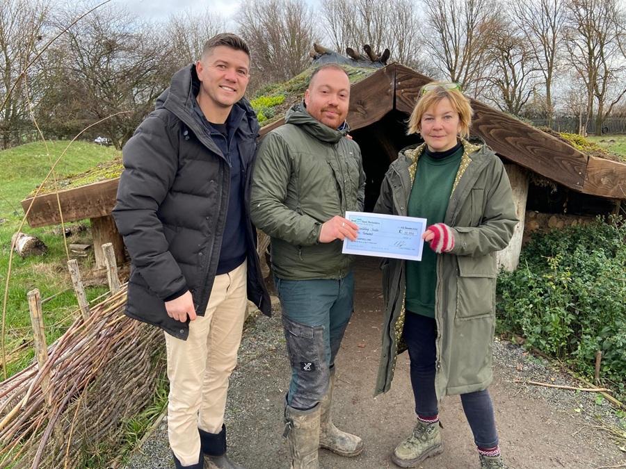 Three people standing outdoors in front of a rustic wooden shelter. The person in the middle is holding a cheque, while the two others stand on either side smiling.