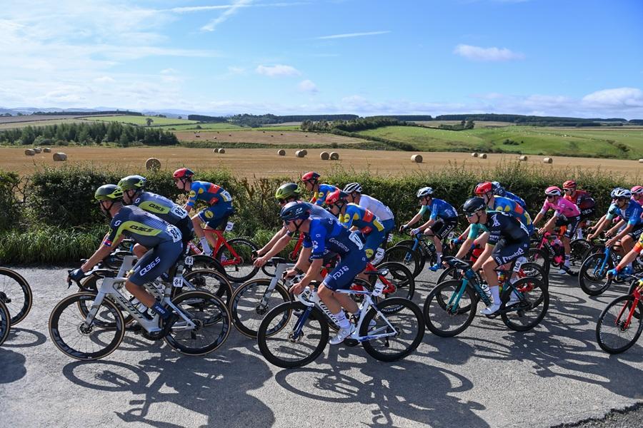 A peloton of cyclists races along a country road, with fields and hay bales under a blue sky.