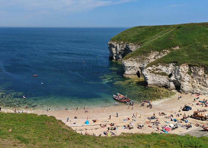 A coastal beach scene with people sunbathing, swimming, and relaxing on a sandy shore beneath white chalk cliffs covered in green grass. A small boat is pulled up near the water, and the sea is calm and deep blue under a bright sky.