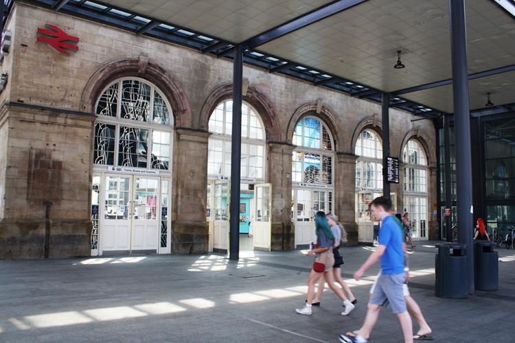 Exterior concourse of Hull Paragon Interchange with arched stone windows and a modern canopy overhead, as several pedestrians walk across the open paved area in daylight.