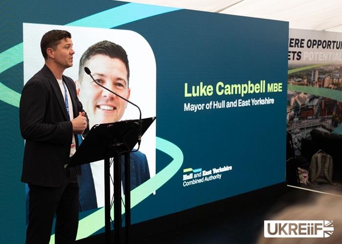 Mayor Luke Campbell speaking at a podium in front of a large presentation screen at a business event.