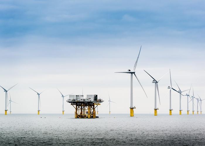 A wide shot of an offshore wind farm in the ocean under a cloudy sky, featuring a central yellow transformer platform surrounded by several large white wind turbines.